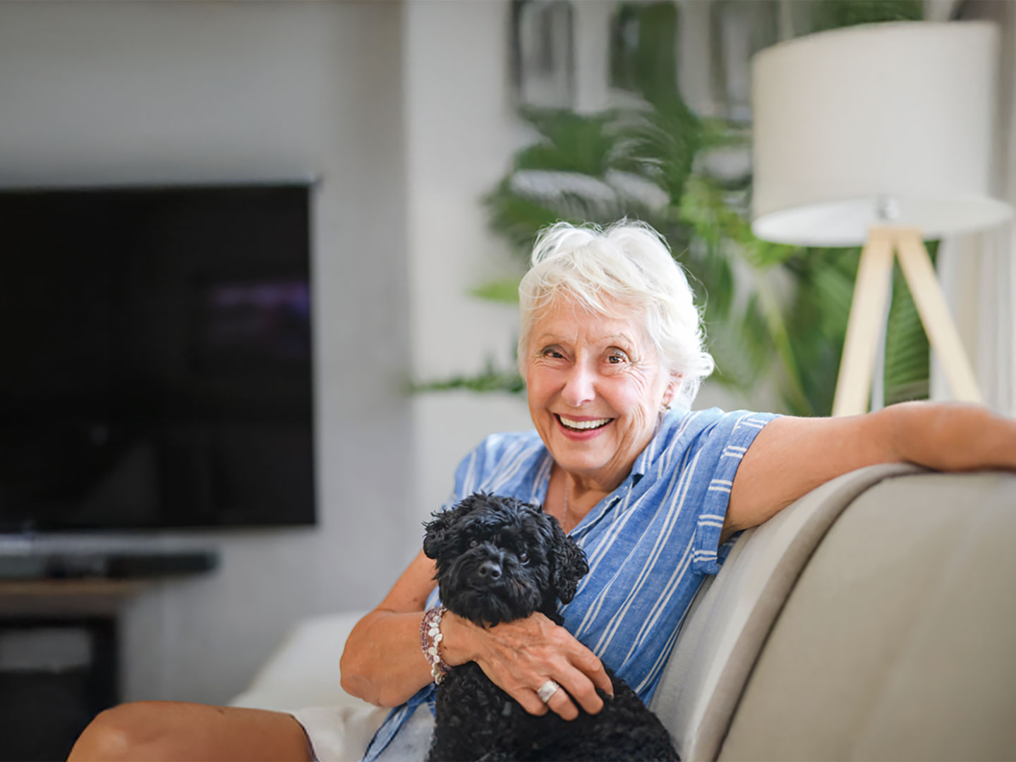 A woman in her seventies, smiling, sitting on a sofa at home with her dog