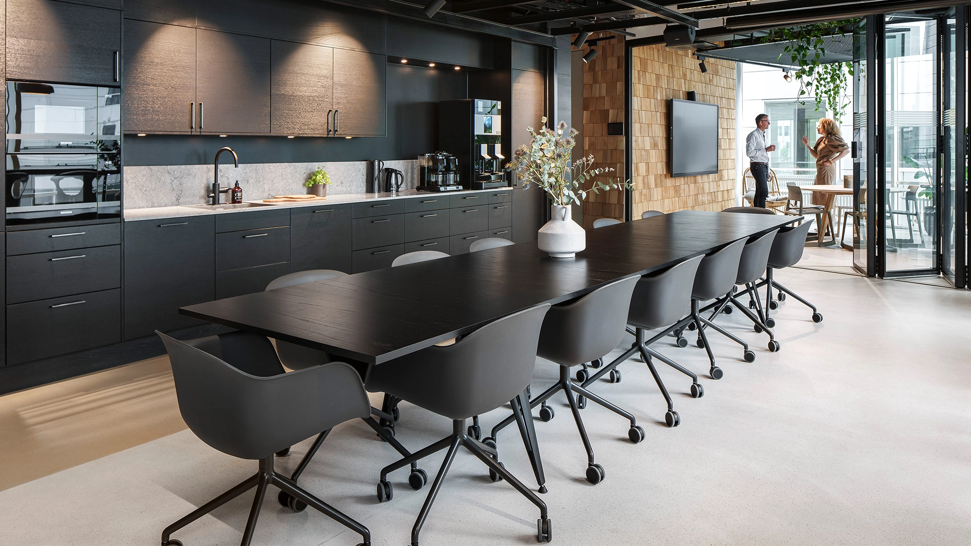 Modernt kontorskök med eleganta svarta möbler, vita bänkskivor och rikligt med naturligt ljus genom glasdörrar och takfönster.