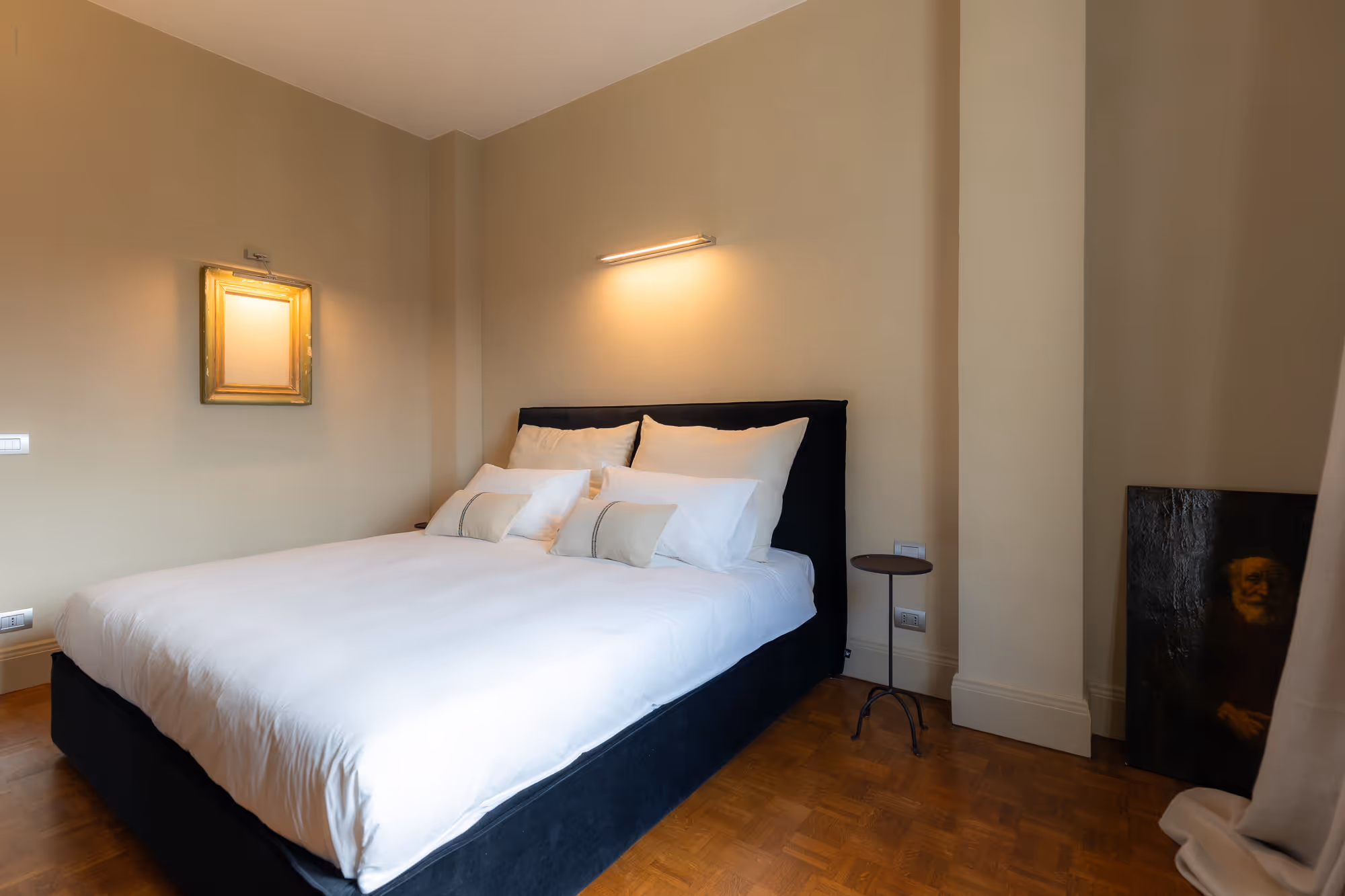 Camera da letto con letto grande nero, lenzuola bianche, cuscini e due quadri alle pareti.