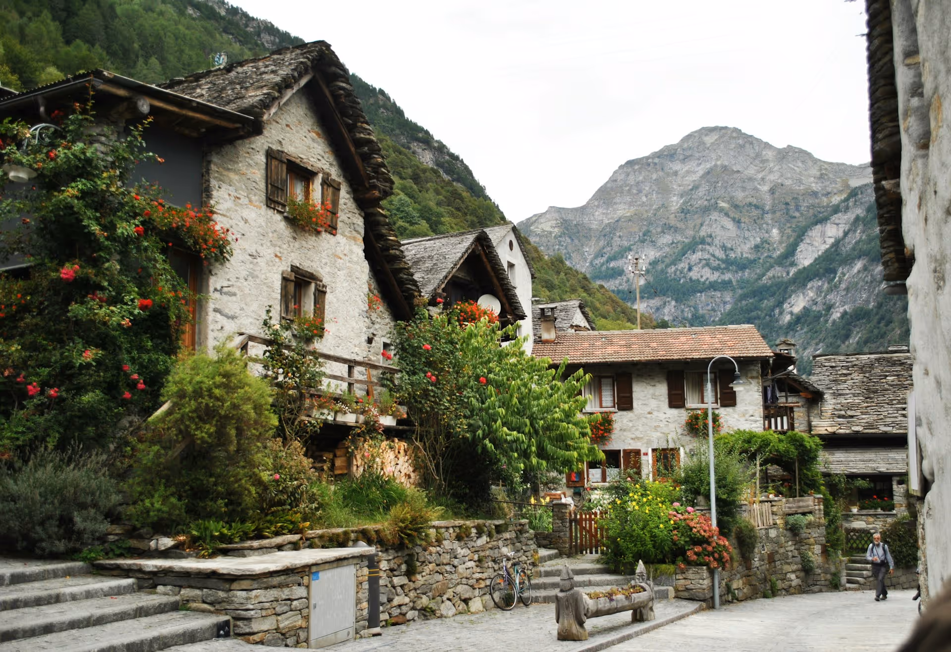 Antico villaggio alpino con case in pietra, fiori rossi e montagne innevate sullo sfondo.