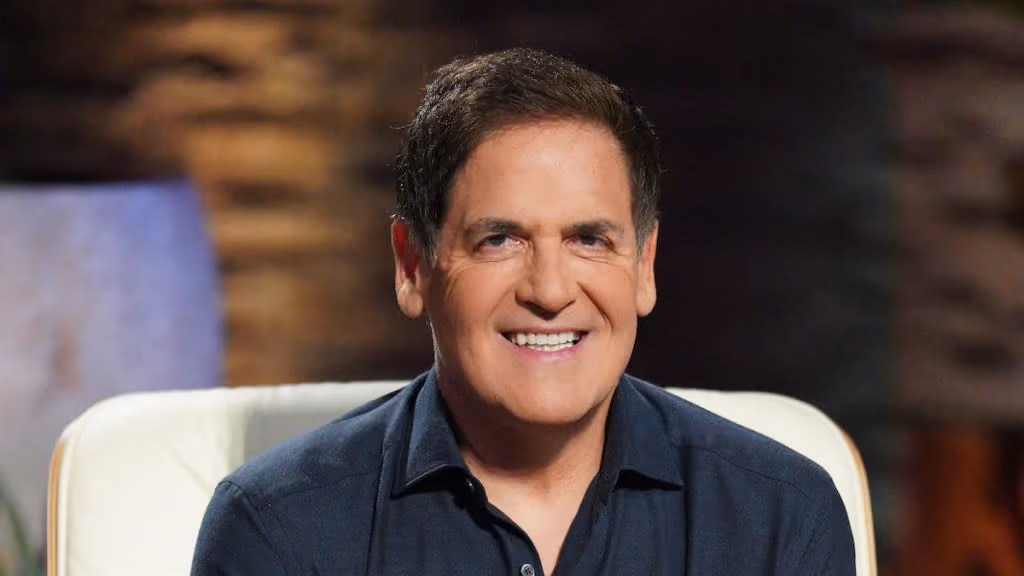 Smiling man with dark hair wearing a dark blue shirt sitting on a white chair with a blurred background.