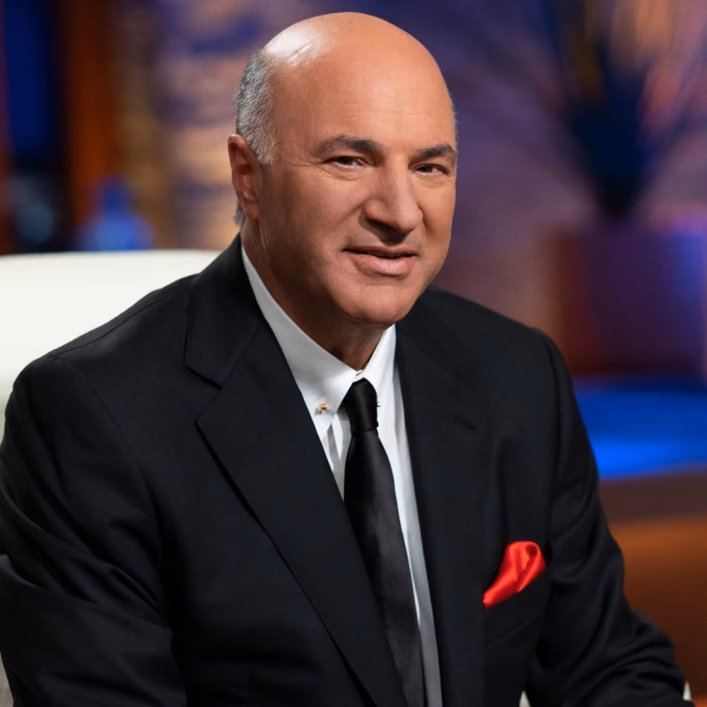 Man in black suit with white shirt and red pocket square smiling with blurred background.