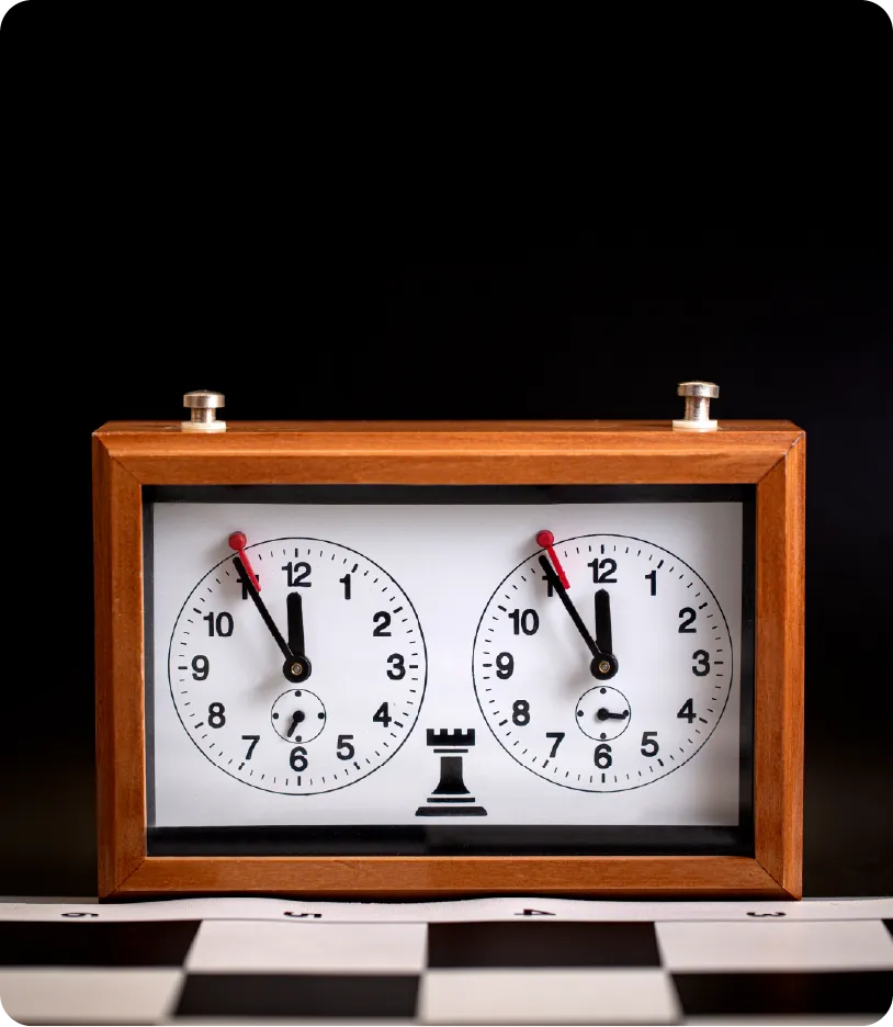 A wooden chess clock with two round white dials, each showing a time of slightly over 6 minutes, placed against a black background. The clock features a chess piece graphic at the bottom.