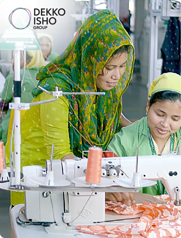 Two women in a garment factory, one wearing a green headscarf and the other in a green uniform, working together on a sewing machine.