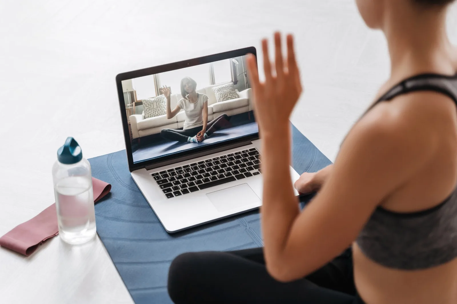 Person on yoga mat waving at a woman on a laptop screen during an online fitness session.