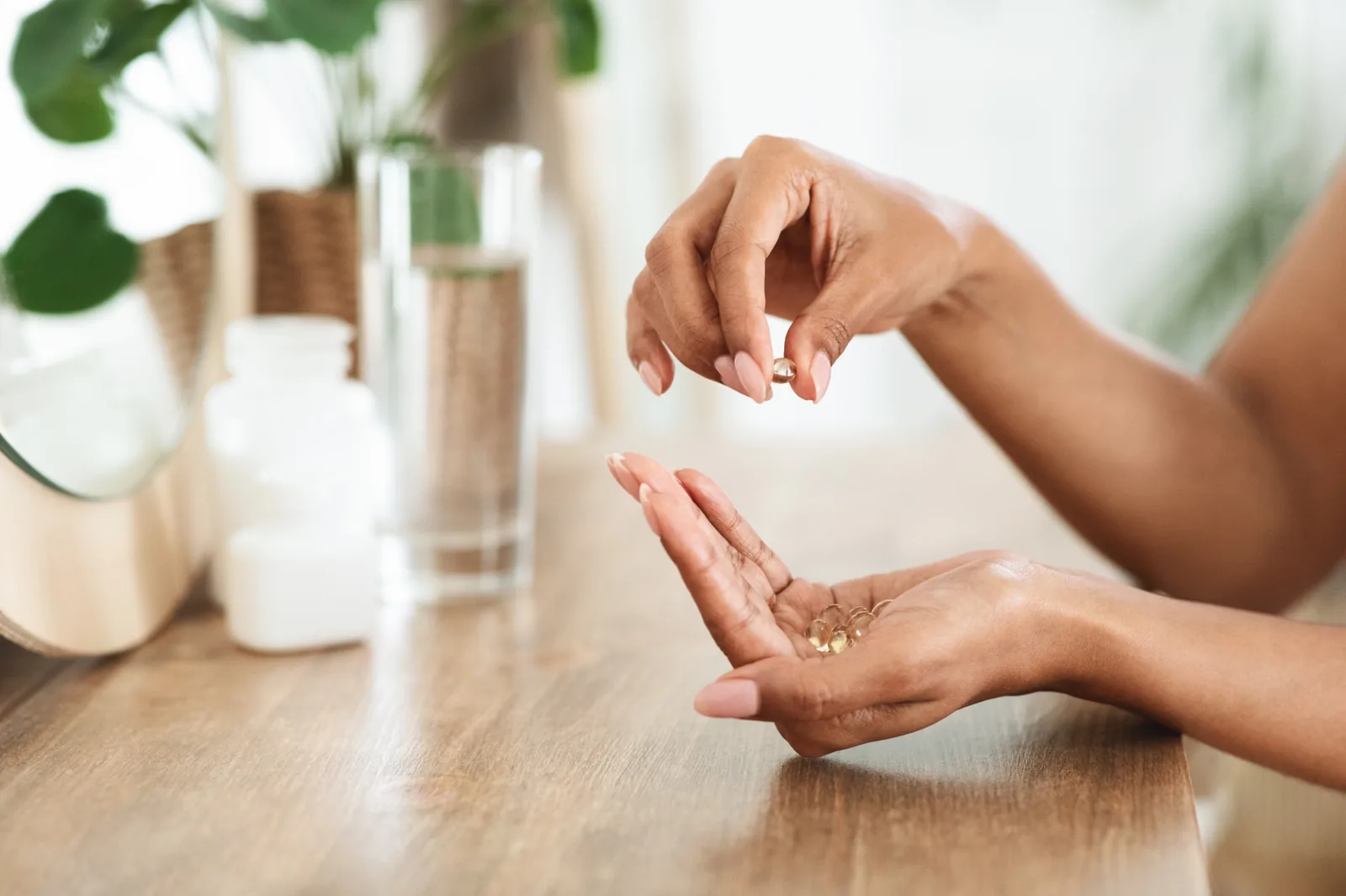 Hands holding translucent supplement capsules over a wooden table with a glass of water nearby.