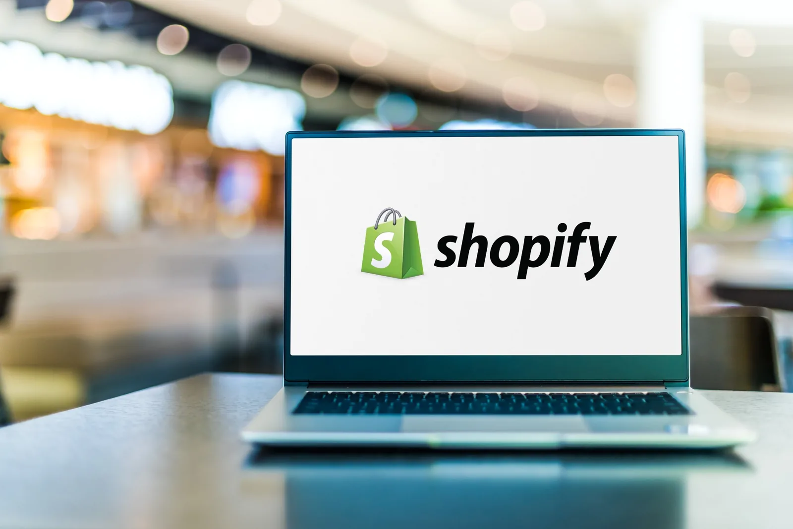 Laptop screen displaying Shopify logo with green shopping bag icon on table in blurred background.