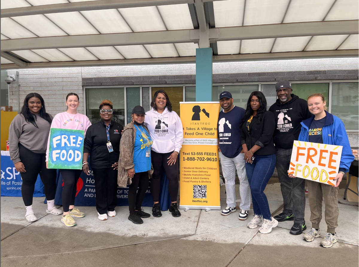 Group of volunteers holding "Free Food" boards standing next to an informative floor banner with information about It Takes A Village To Feed One Child programs