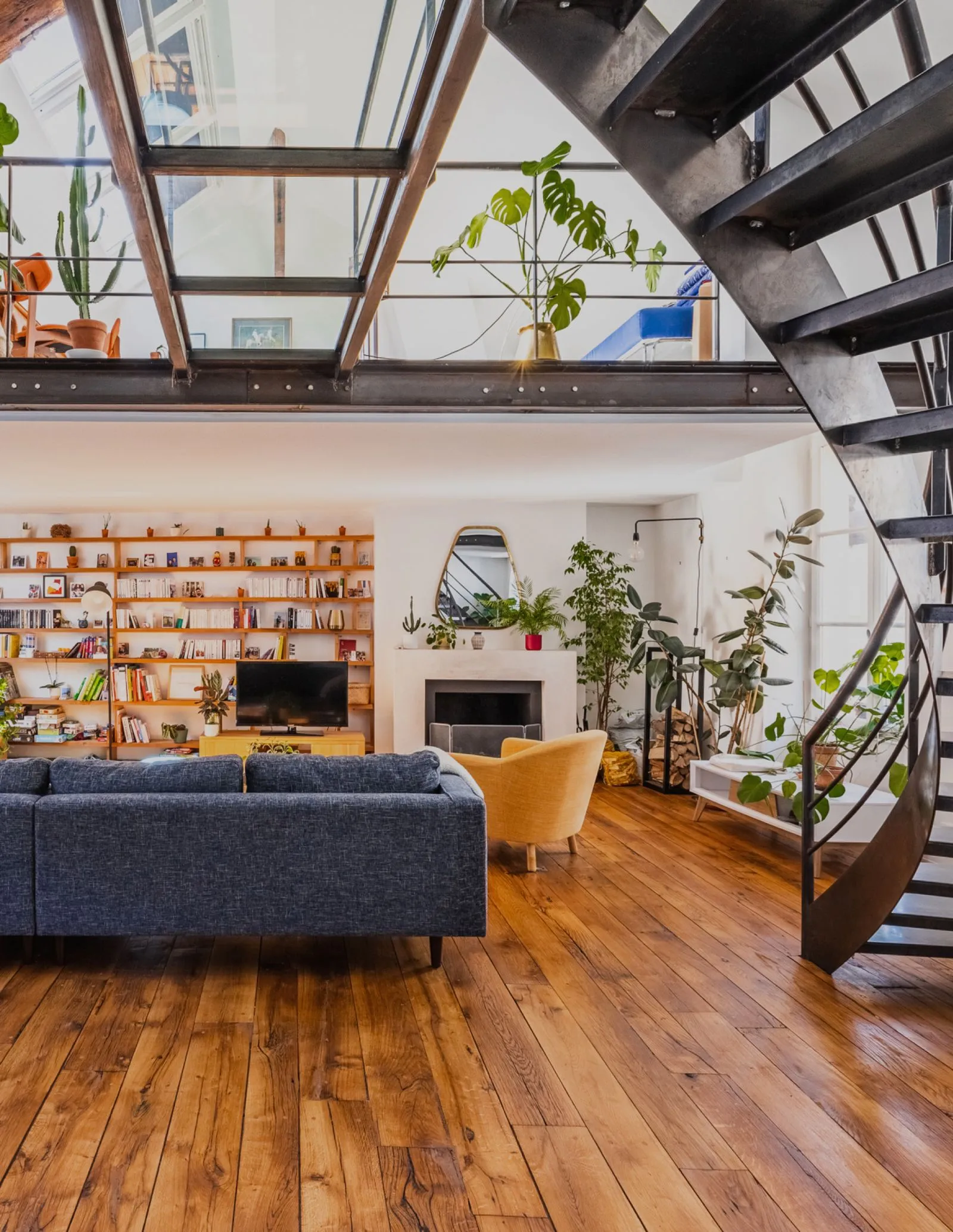 Salon lumineux d'un loft moderne avec un escalier en métal et bois, un grand canapé bleu, une bibliothèque en bois et de nombreuses plantes.