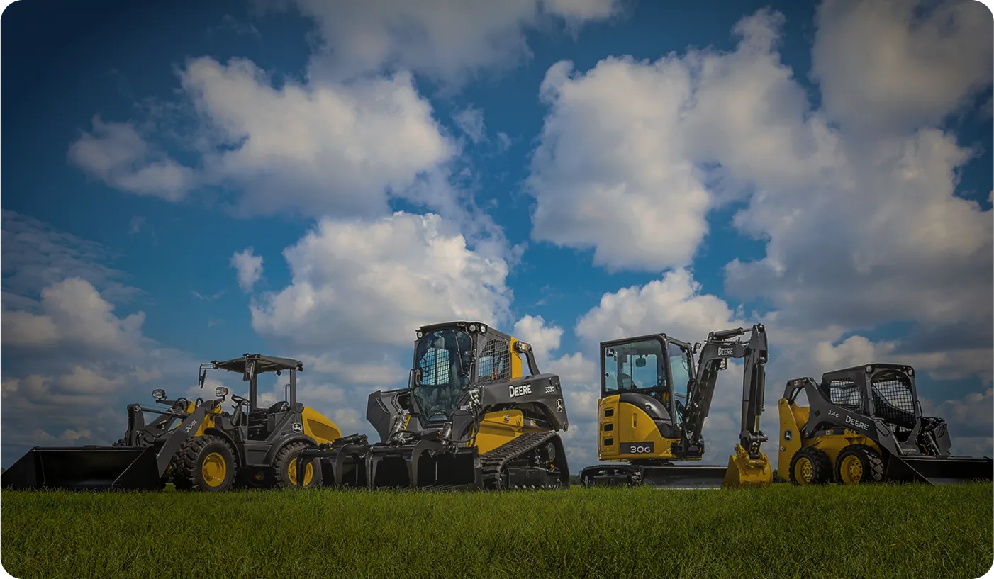 Four yellow and black John Deere construction vehicles lined up on green grass under a blue sky with white clouds.