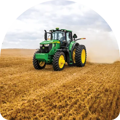 Green tractor with yellow wheels driving across a dry harvested field under a cloudy sky.