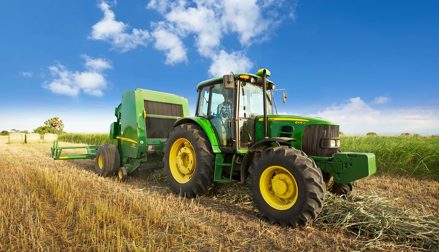 Green John Deere tractor with large yellow wheels operating in a harvested field under a blue sky with scattered clouds.