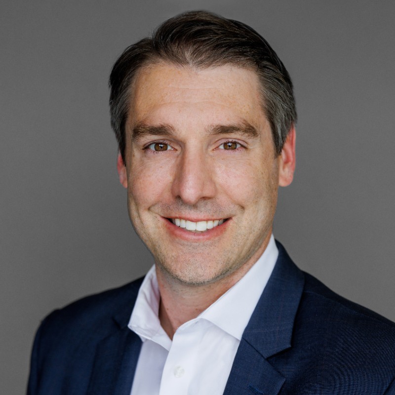 Smiling man with brown hair wearing a navy blue suit and white shirt on gray background.