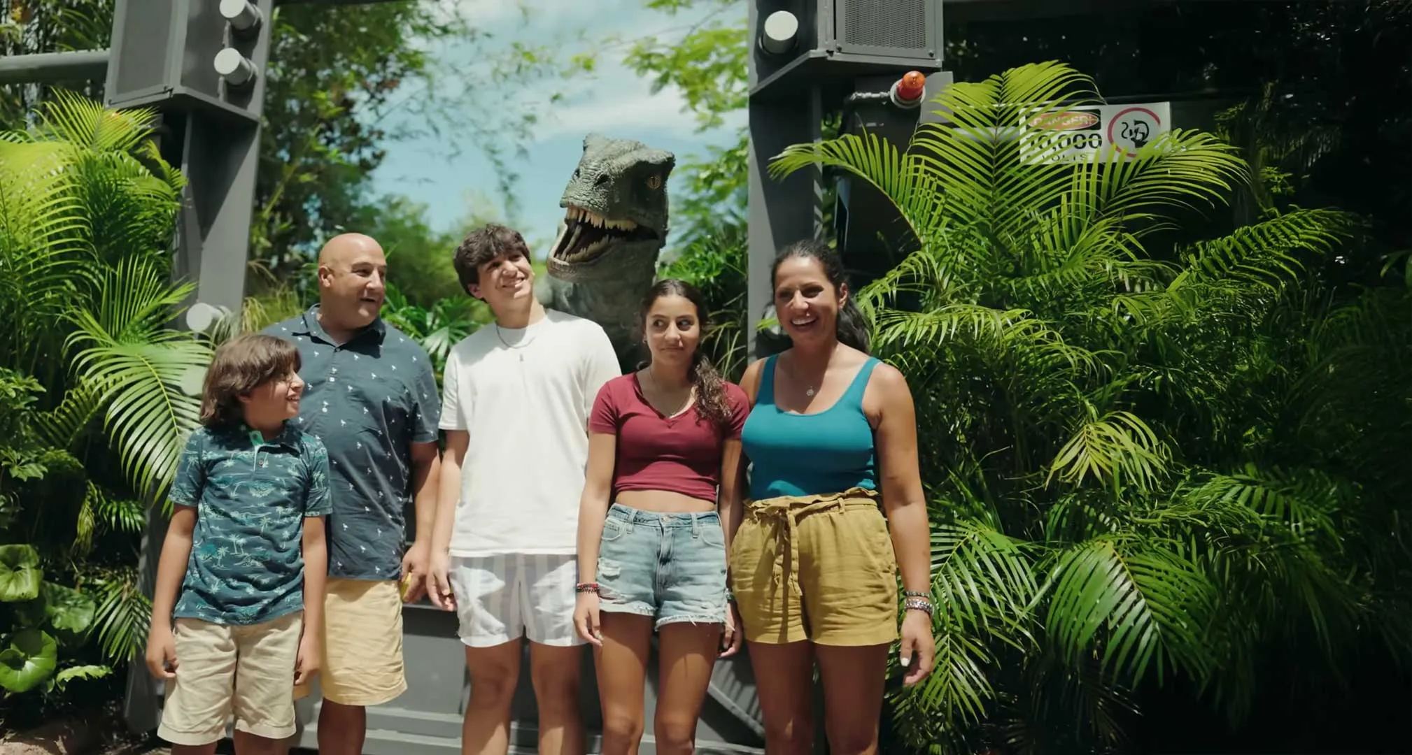 A family of five posing outdoors with a realistic dinosaur model behind them, surrounded by lush green plants.