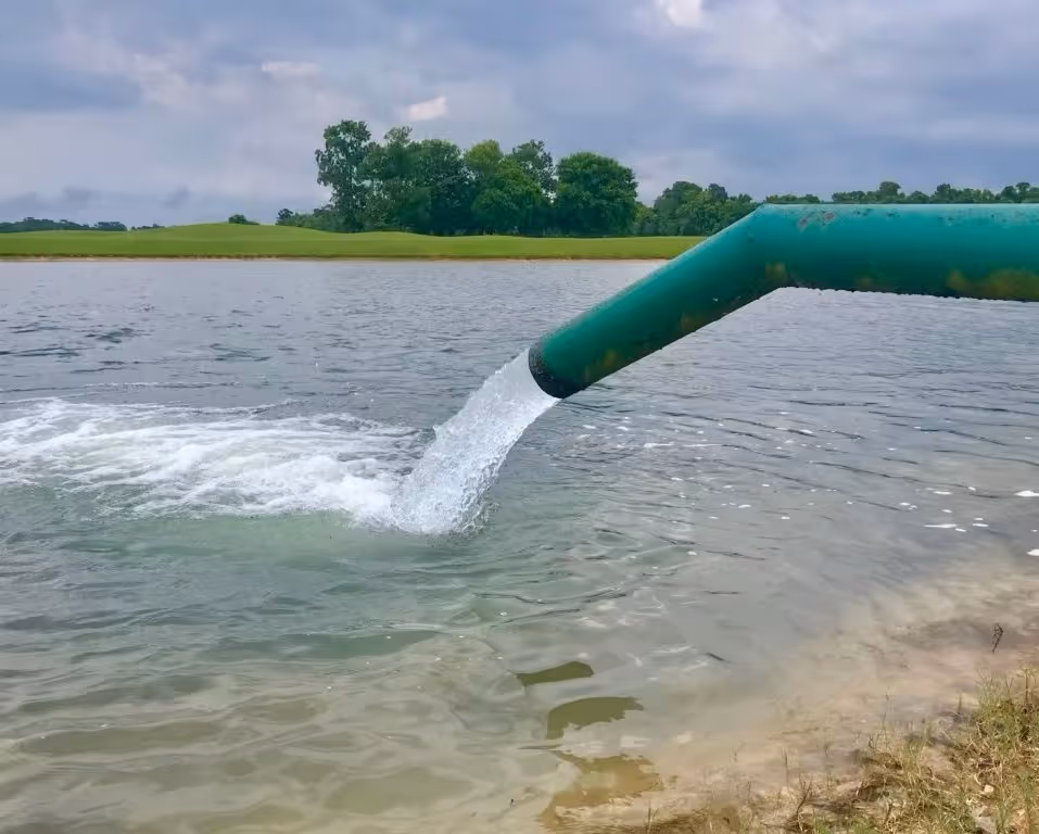 Green pipe discharging water into lake with grassy landscape in background