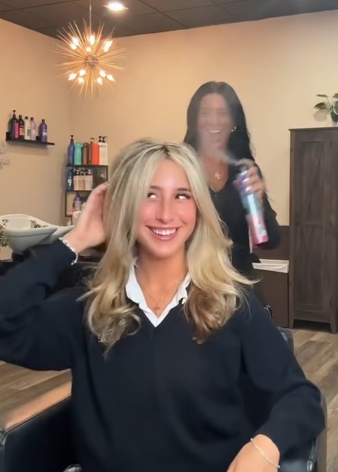A smiling client with freshly styled blonde hair sits in a salon chair while a stylist finishes the look with hairspray at Razberris Hair, Nail & Spa Salon in Toms River, NJ.