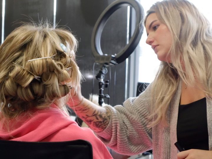 Ashley, hairstylist and makeup artist at Razberris Hair, Nail & Spa Salon in Toms River, applies makeup to a client with hair rollers under a ring light during a salon beauty session.