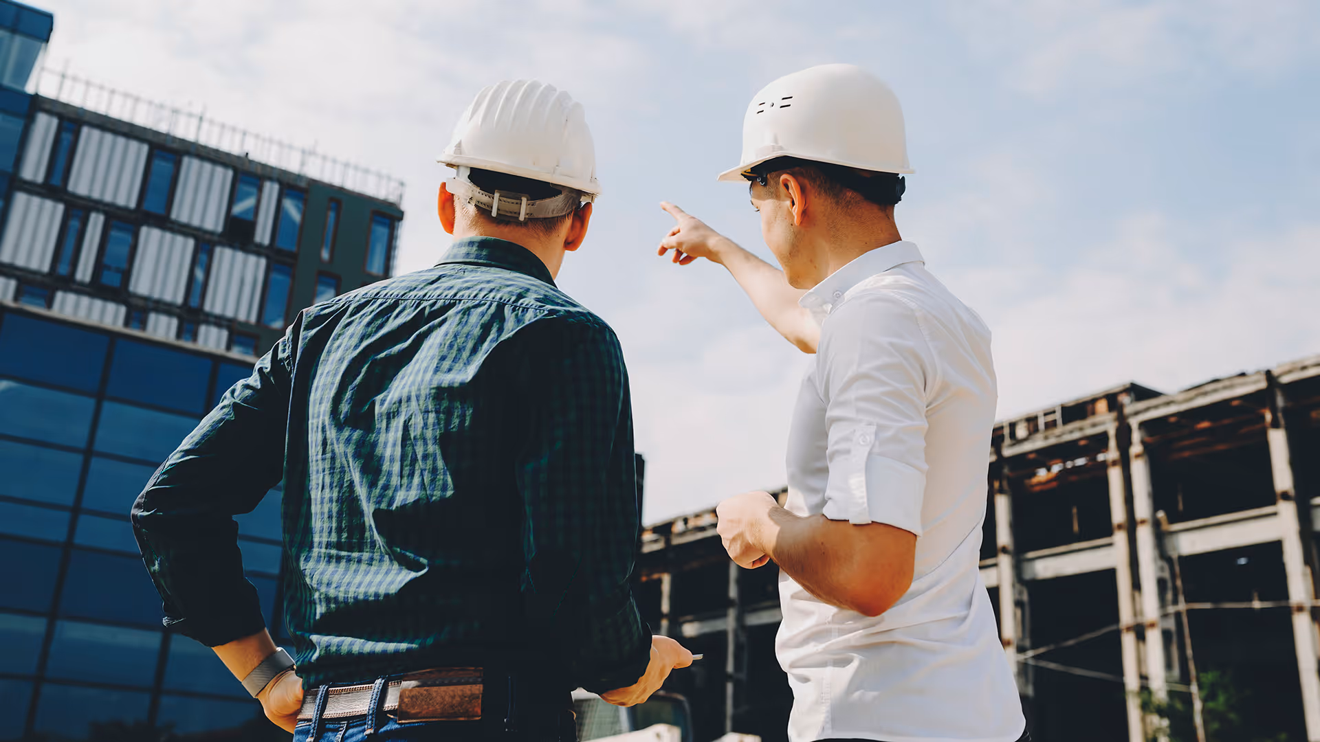 Two construction workers in hard hats discussing progress while pointing at a modern building site.