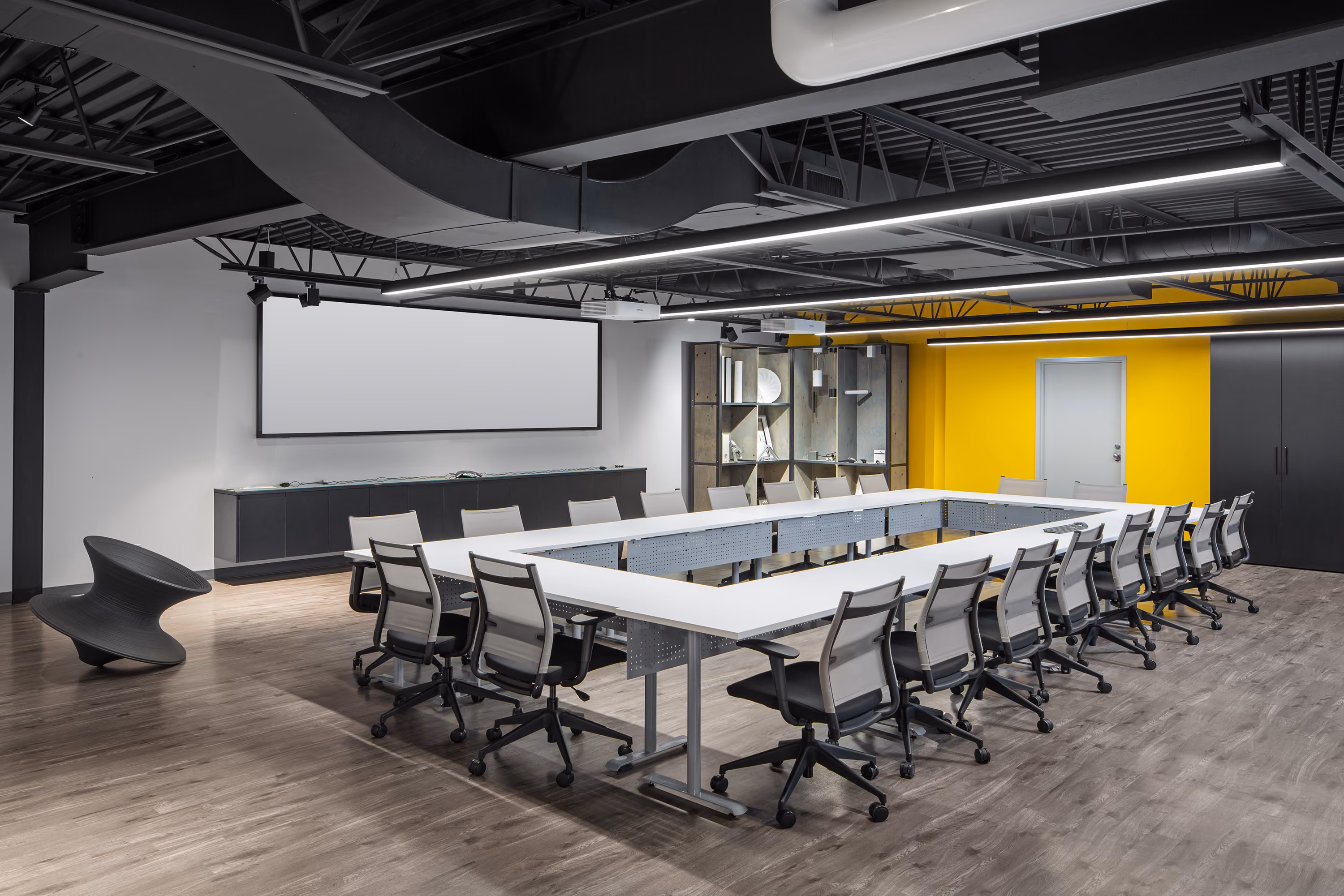 Modern conference room with a large U-shaped white table surrounded by mesh office chairs, a blank projector screen, yellow accent wall, and wood flooring.
