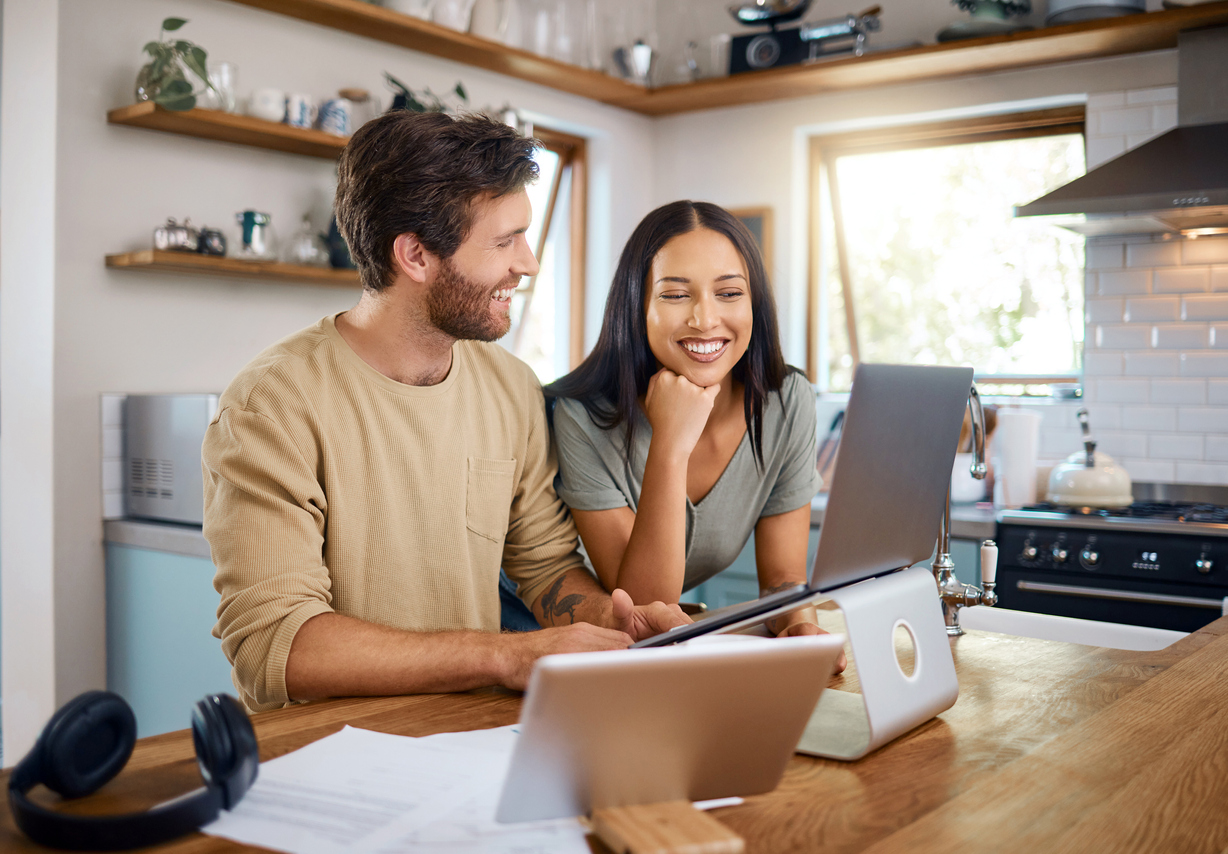 Happy young caucasian man working on laptop while his wife stands next to him looking at the screen. A black wallet with credit cards.