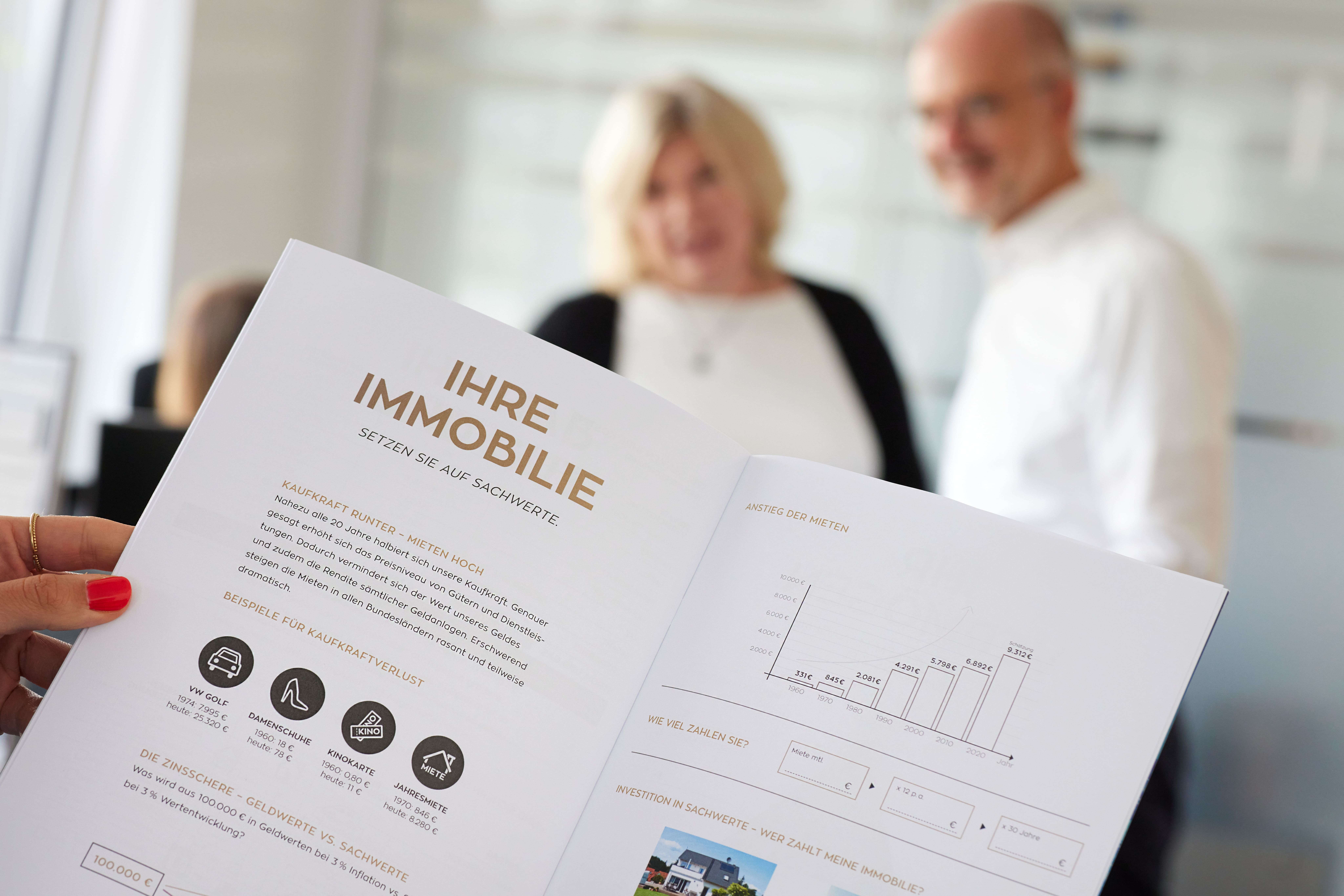 Beratungsunterlage zum Thema Immobilien mit der Aufschrift ‚IHRE IMMOBILIE – Setzen Sie auf Sicherheit‘; im Hintergrund zwei verschwommen erkennbare Berater – symbolisch für ganzheitliche Finanzberatung mit persönlicher Begleitung.