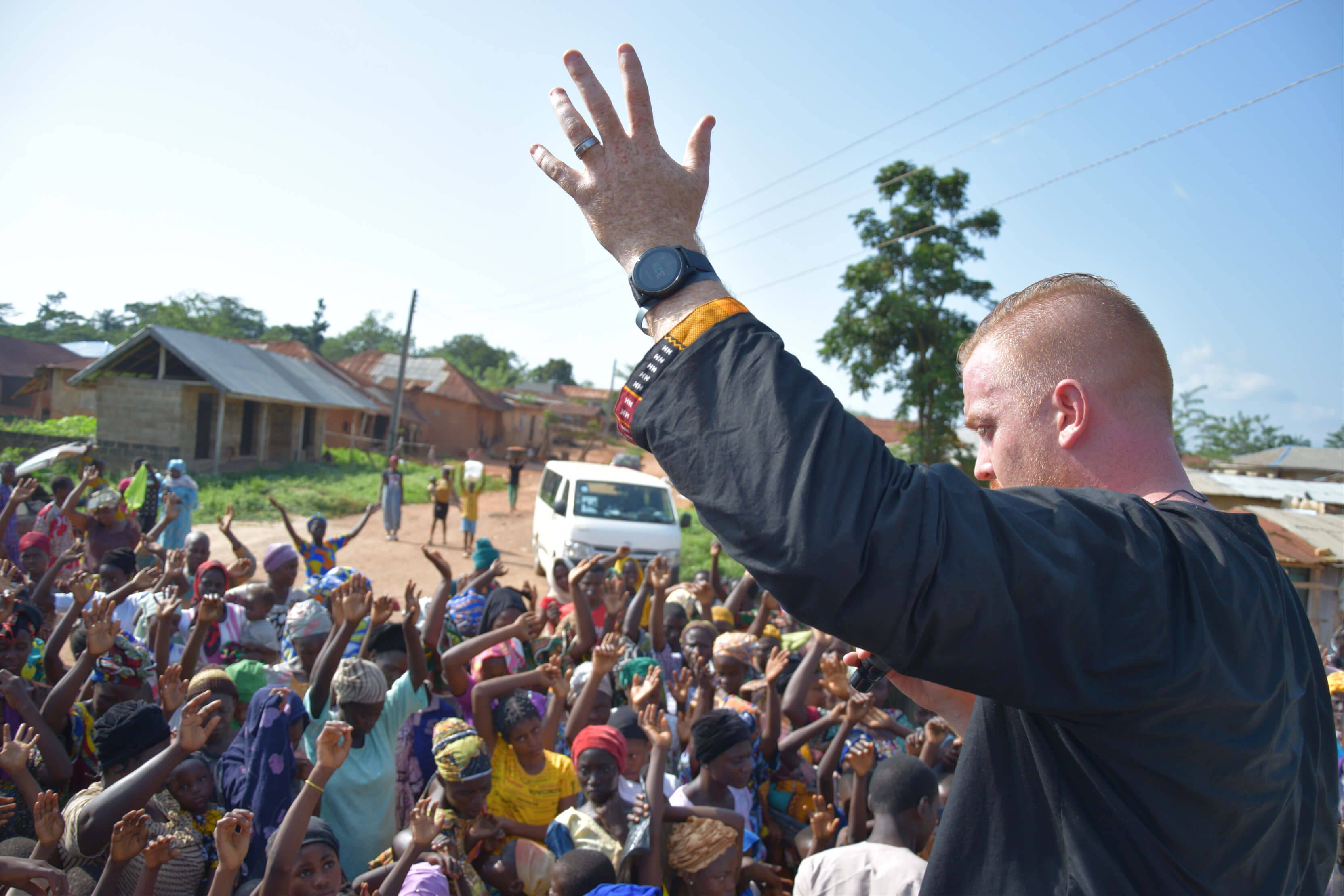 A crowd stands with their hands raised high, radiating passion and energy in a village setting. Andrew raises his own hands in a moment of sacred proclamation.