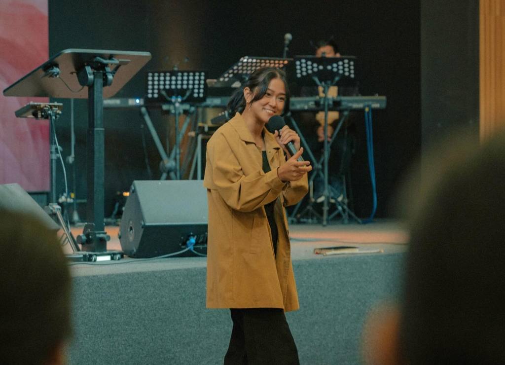 Anne stands on the ground, holding a microphone and speaking passionately to the audience about Jesus. Behind her, a stage serves as the backdrop. She addresses the crowd at eye level, creating a sense of connection and intimacy.