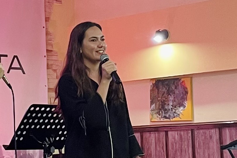 Kathy stands before the crowd, microphone in hand, her voice filled with passion. Behind her, a music set is arranged alongside a picture frame and a light, creating a warm and inviting backdrop. Her gaze radiates intensity as she connects with her audience.