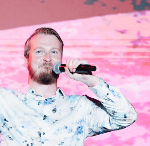 Niki stands against a pink background, holding a microphone and speaking passionately about Jesus.