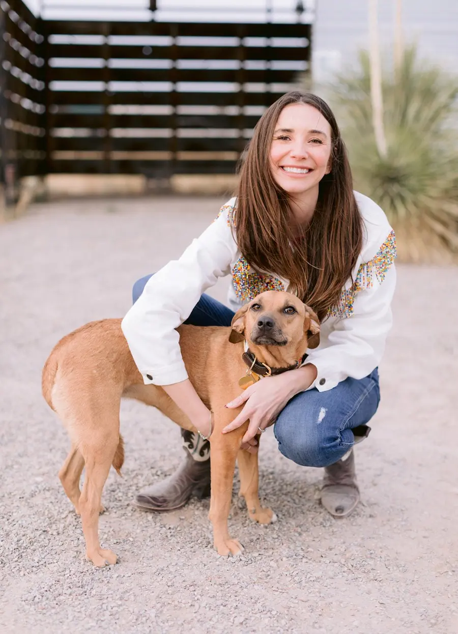 Blaine in a white jacket with colorful fringe, crouching, holding her dog Dee Dee