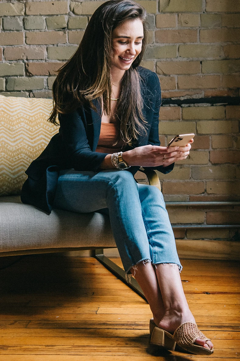 Blaine seated on a grey couch in a brick loft, looking at her phone in her hands, and smiling
