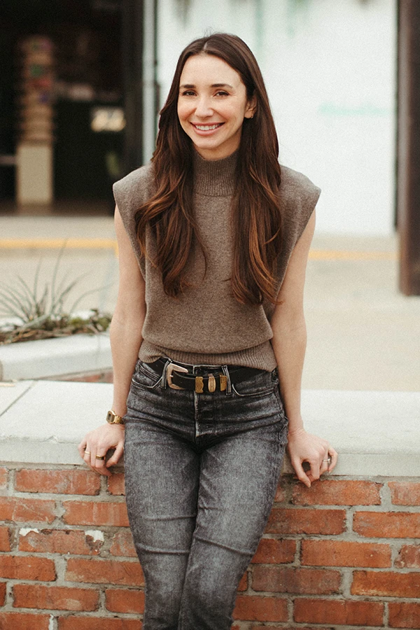 Blaine in a tan top seated on a brick wall facing the camera, smiling