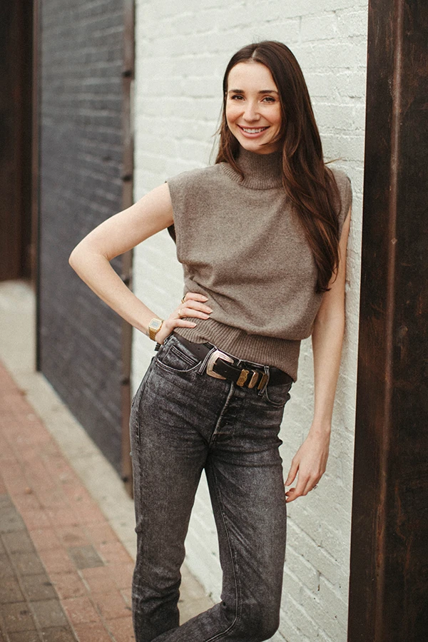 Blaine in a tan top, leaning against a white brick wall, smiling