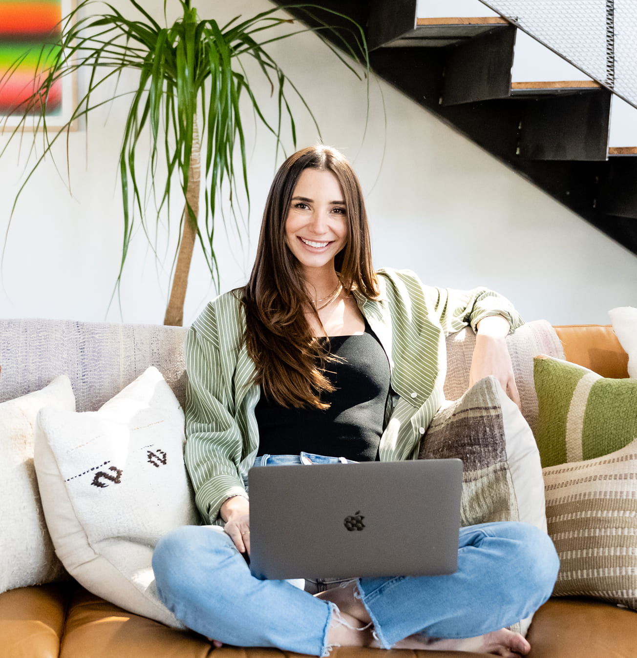 Smiling woman sitting cross-legged on a couch with pillows, using a laptop, with a plant and staircase in the background.