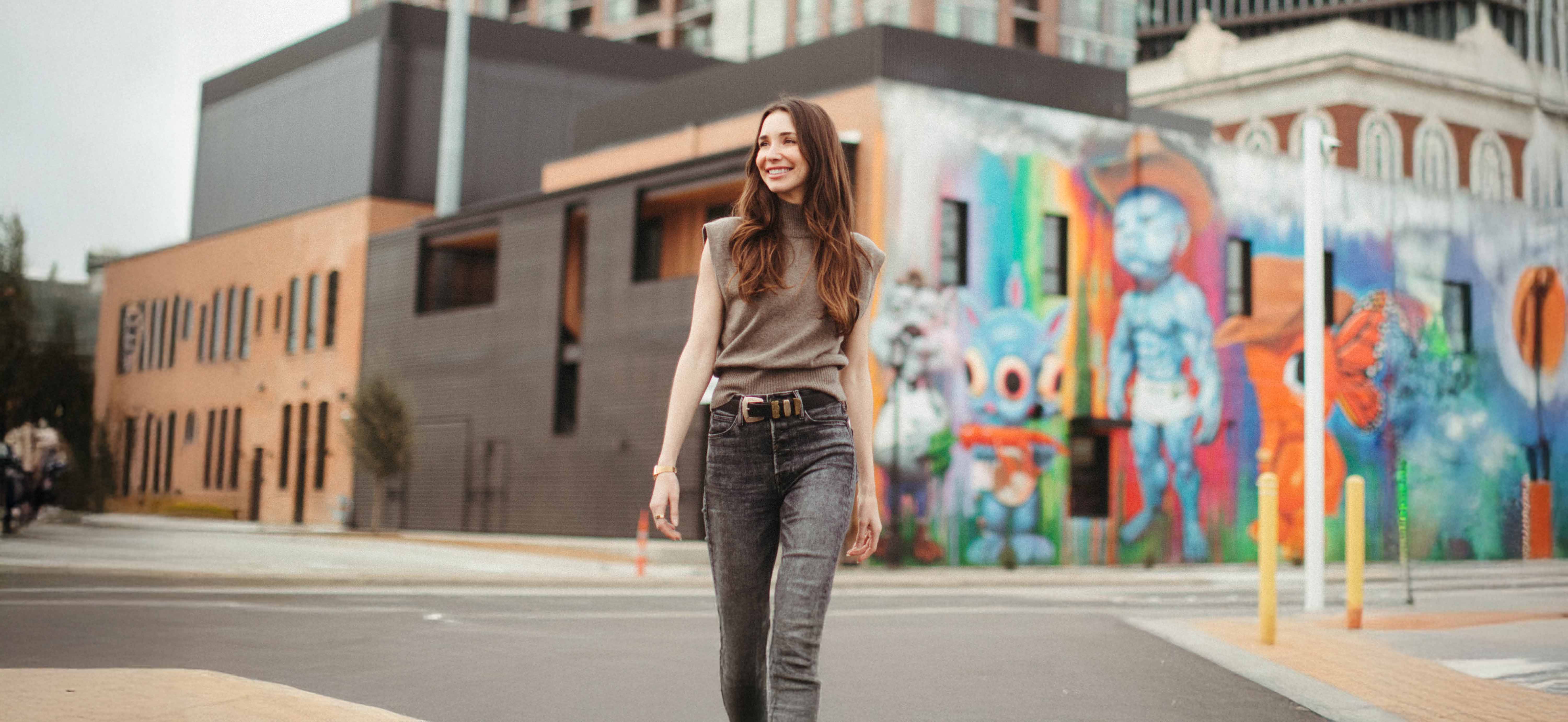 Blaine, smiling, walking on a city street with colorful mural artwork on buildings in the background.