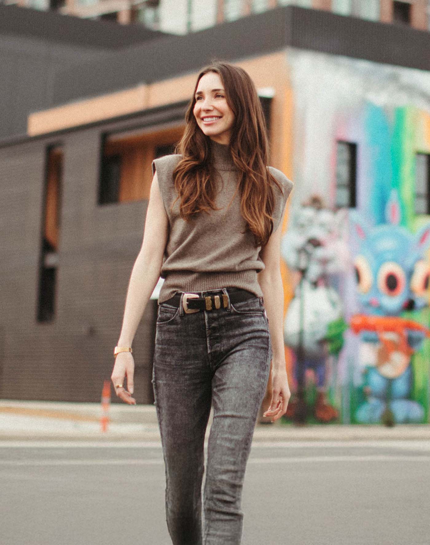 Blaine wearing a sleeveless gray top and dark jeans walking on a city street with colorful graffiti in the background.