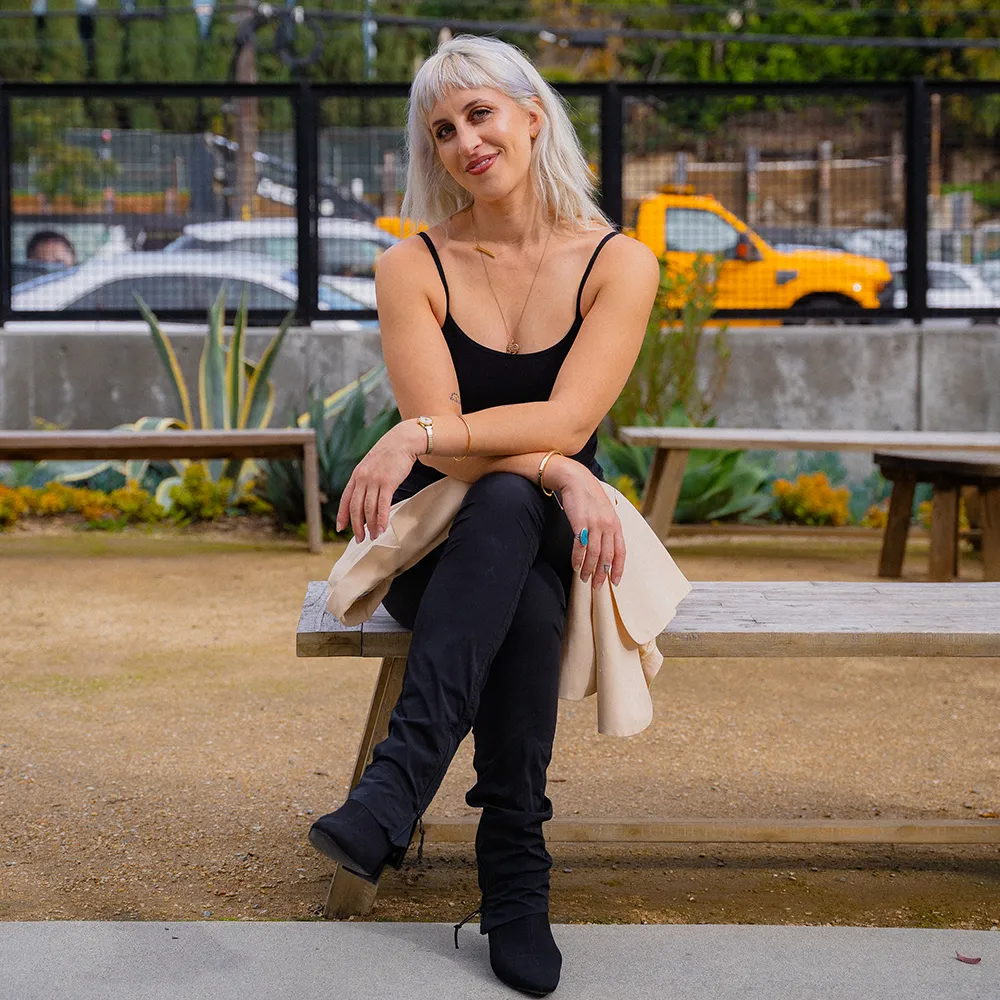 Photo of Merav seated, in a black tank top and black jeans, smiling