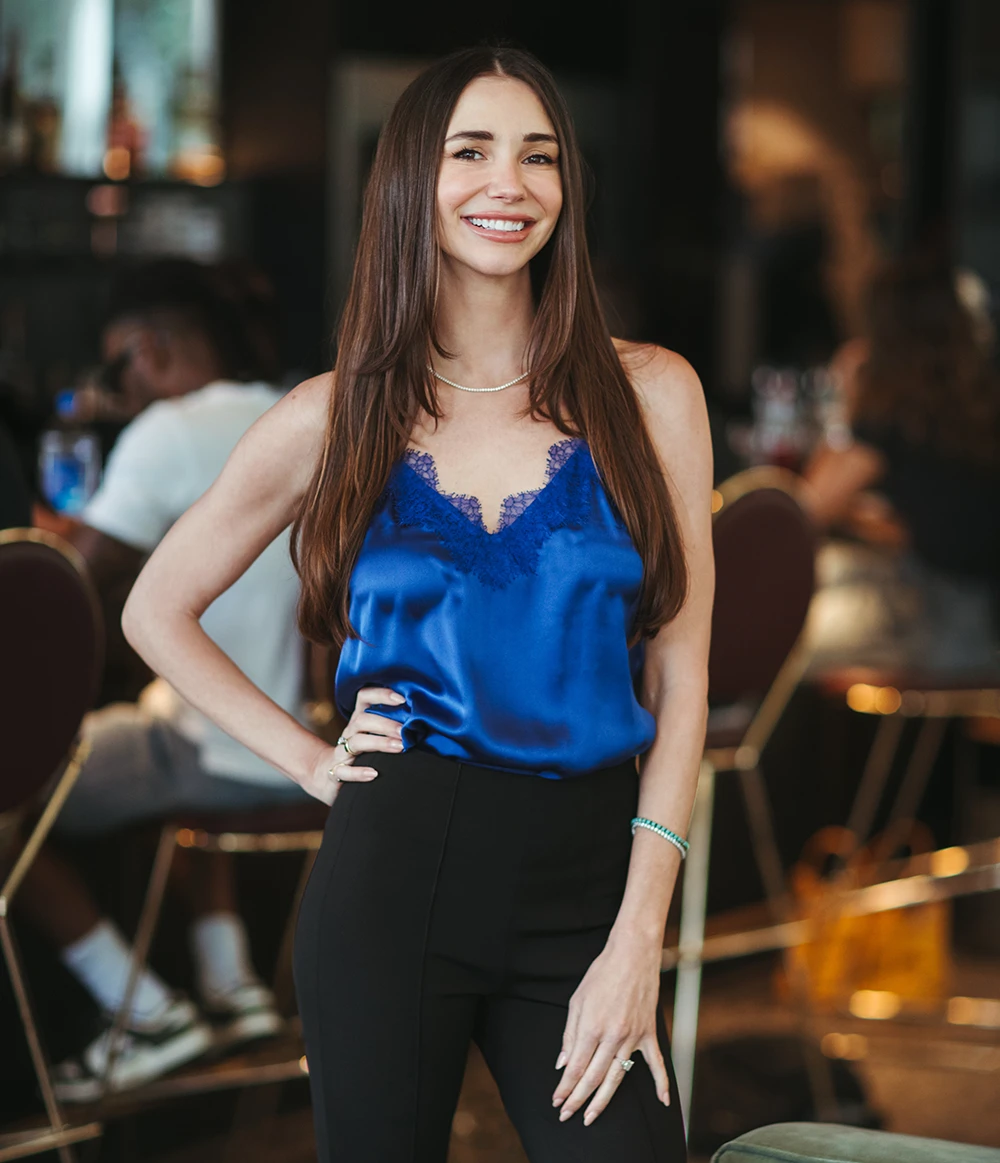 Blaine, wearing a white button-up shirt and beige skirt, standing indoors, smiling.