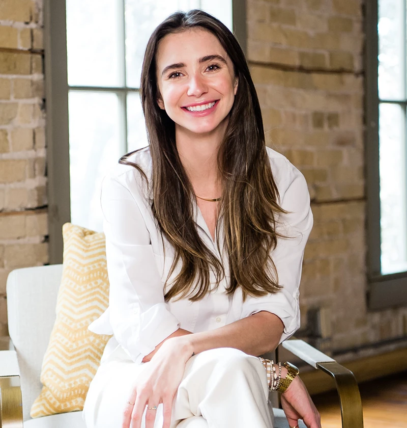 Blaine, wearing a white button-up shirt and beige skirt, standing indoors, smiling.