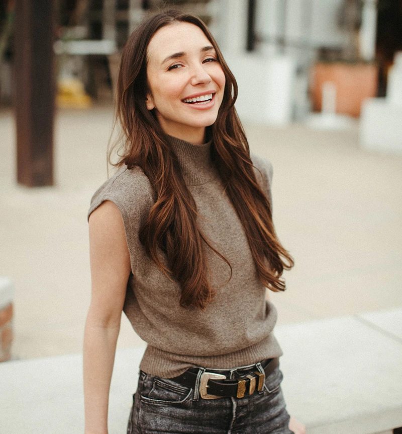 Blaine, wearing a white button-up shirt and beige skirt, standing indoors, smiling.