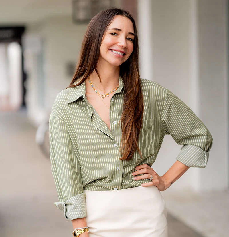 Blaine, wearing a white button-up shirt and beige skirt, standing indoors, smiling.