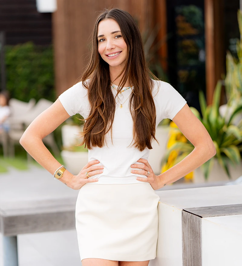 Blaine, wearing a white button-up shirt and beige skirt, standing indoors, smiling.