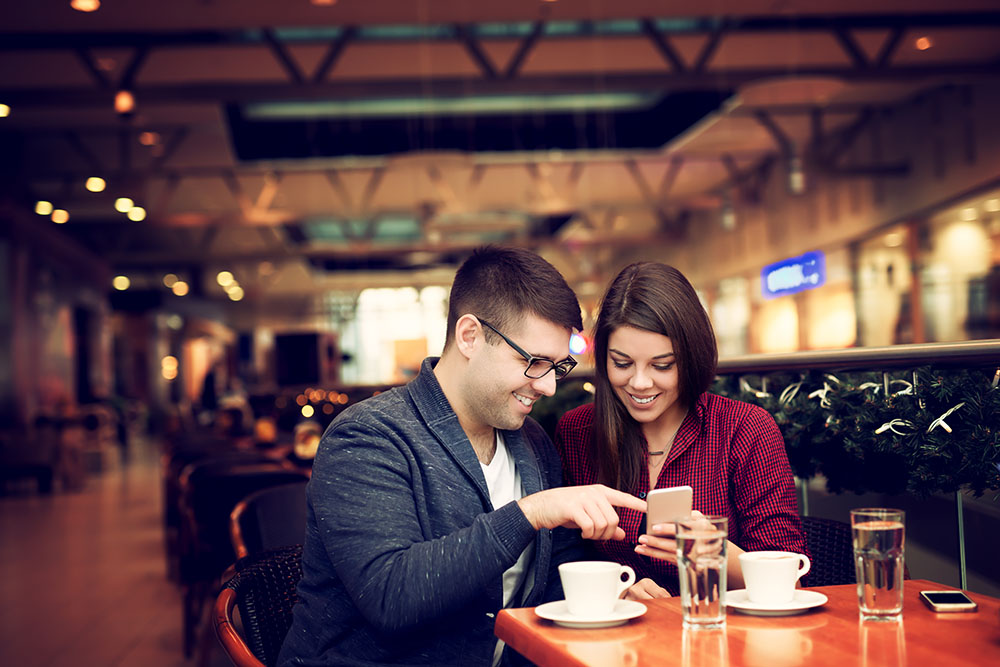 Man and woman seated at a coffee table, both smiling, where man is pointing at a phone the woman is holding