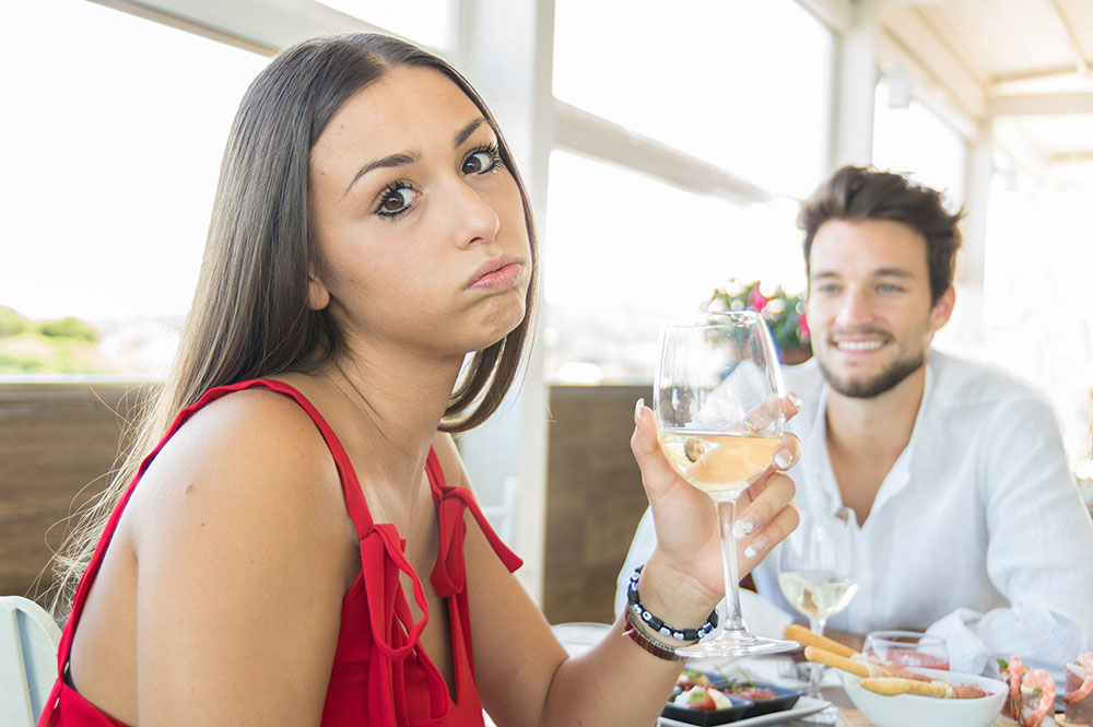 Woman looking into camera with distressed expression, seated at a table with a man, seemingly on a bad date