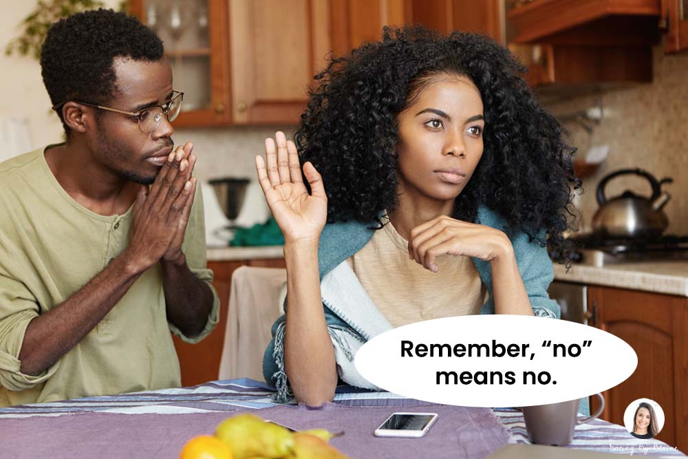 Man with hands in a prayer position, clearly pleading with a woman who stands to his right, and looks disinterested