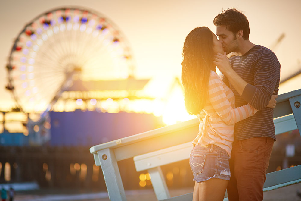 Couple kissing in front of a sunset, with a ferris wheel in the background