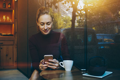 Woman with blond hair in black sweater looking at her phone with a soft smile in a coffee shop