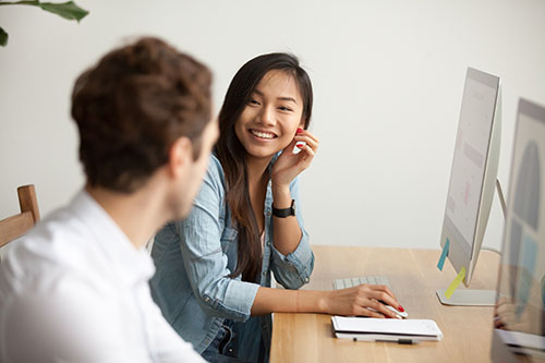 Asian woman seated at a desk, looking to her side and smiling at an unidentified male coworker in the foreground