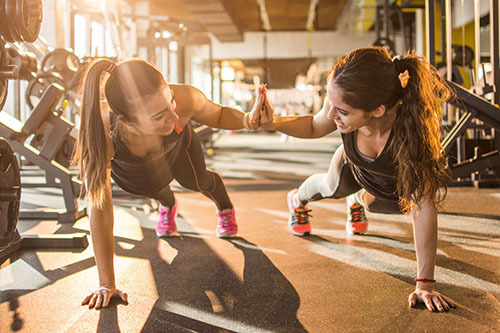 Two brunette women in a gym in one-handed plank position, high-fiving each other while smiling