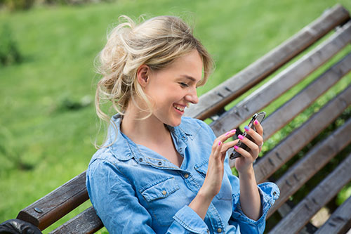 Blond woman seated on a park bench, smiling while looking at her phone screen